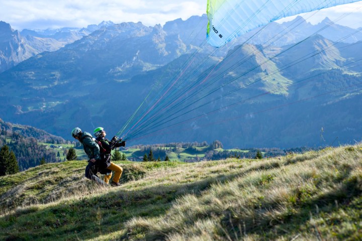 Tandem-Gleitschirmstart auf der Rotenflue: Pilot und Passagier ziehen den Schirm gemeinsam in die Luft. Im Hintergrund eine beeindruckende Berglandschaft mit grünen Hügeln und felsigen Gipfeln.
