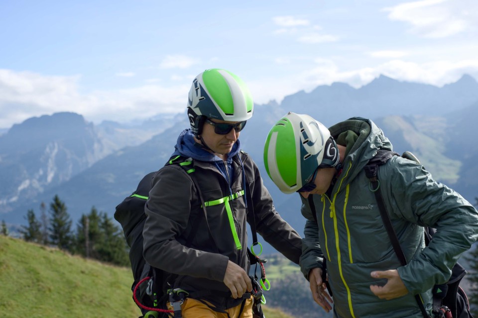Zwei Personen – ein Passagier und ein Tandempilot – mit Helmen und Ausrüstung stehen auf dem grasbewachsenen Hügel der Rotenflue und bereiten sich auf den Gleitschirmflug vor.