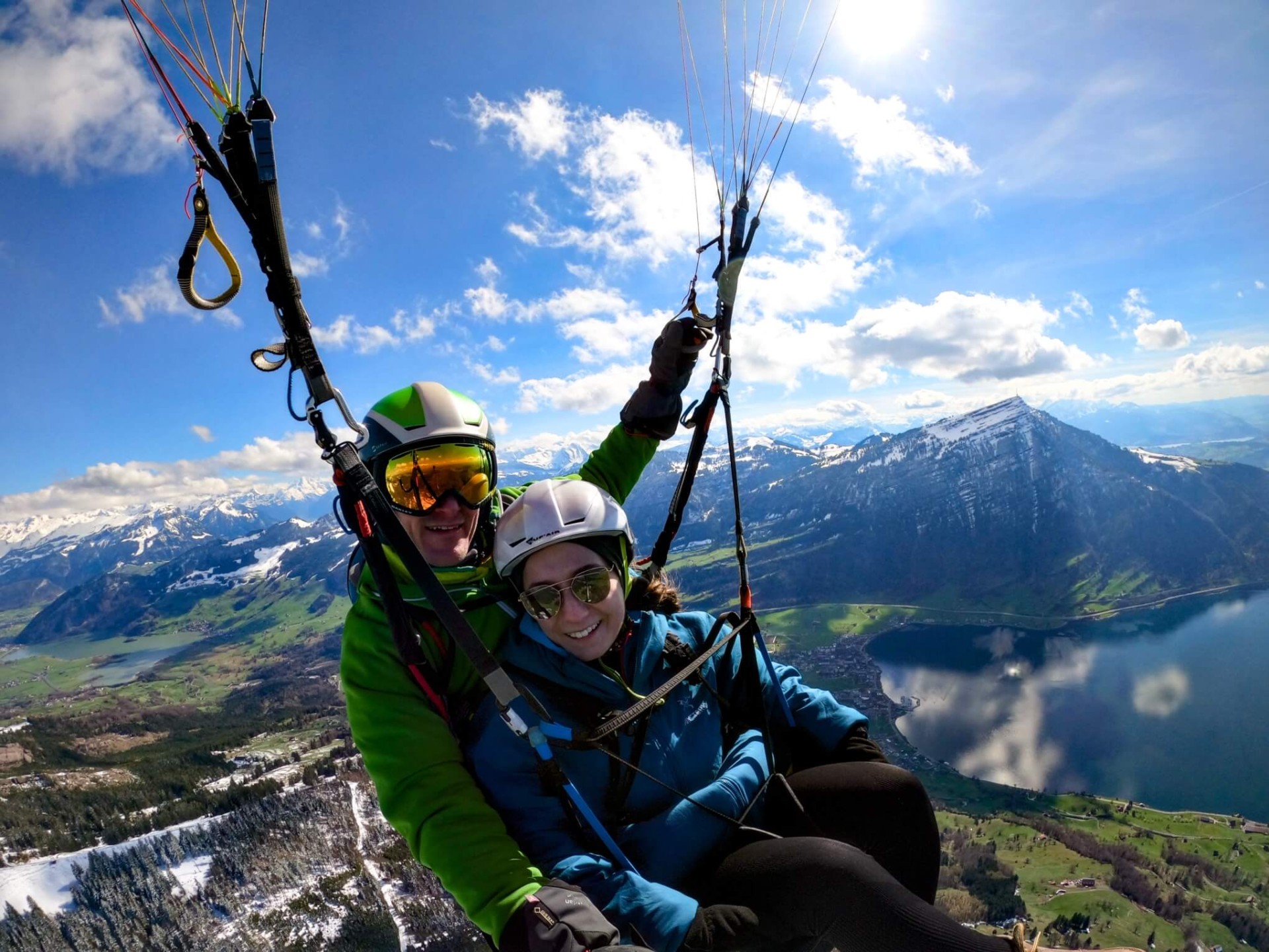 Tandem pilot Pascal with passenger Celina, both laughing, during a paragliding flight over Gnipen. Below them, springtime snowfields and green meadows stretch out. Lake Zug reflects the bright blue sky.