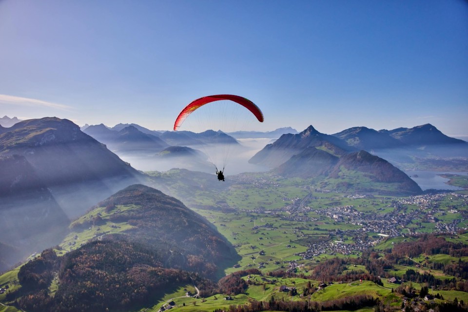 Roter Tandem-Gleitschirm mit Pilot und Passagier unter blauem Himmel, über grünen Feldern, dem Vierwaldstättersee und den Bergen Rigi-Kulm, Rigi-Scheidegg und Urmiberg.