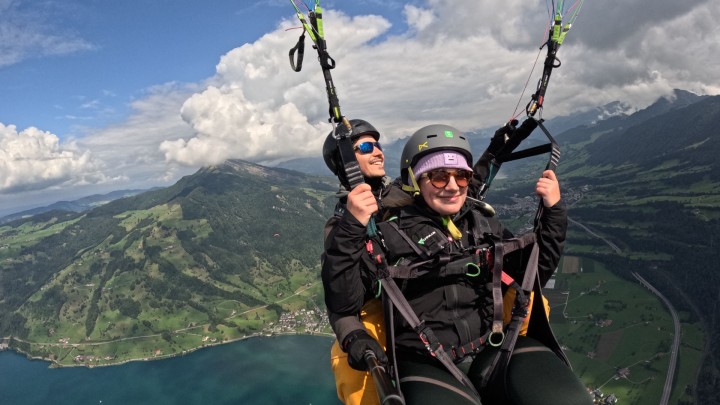Ein Tandem-Gleitschirmflug mit Pilot Daro Flückiger und strahlender Passagierin über dem Zugersee. Im Hintergrund erstrecken sich grüne Hügel, dichte Wälder und der markante Gipfel des Rossbergs.