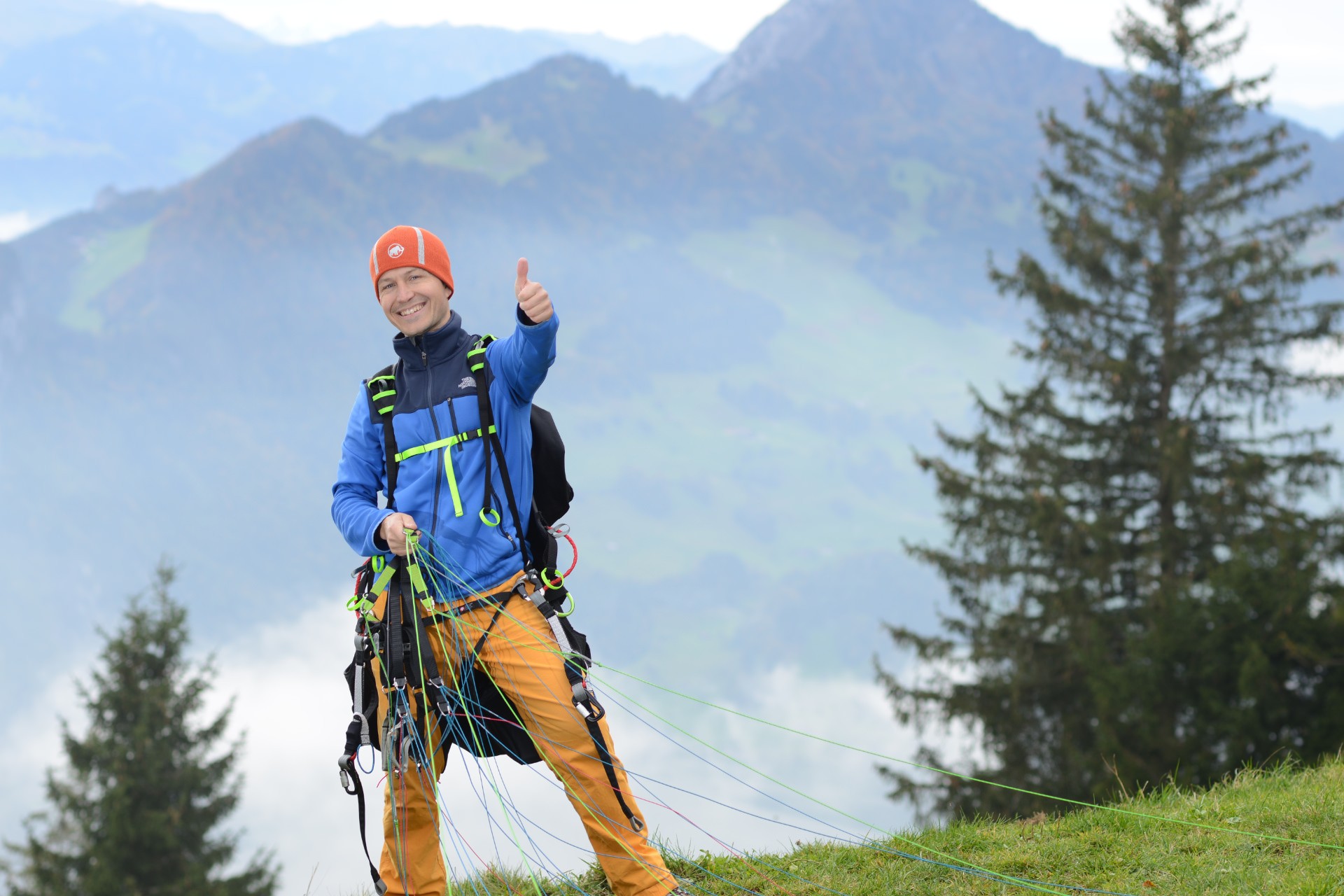 Tandem pilot Daro gives a thumbs-up at the launch site, signaling his excitement. He is wearing a paragliding harness and holding the colorful lines of the canopy. In the background: Rigi Hochflue.