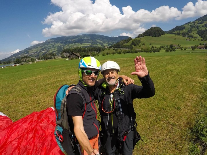 Tandempilot Pascal und Passagier Oskar stehen lachend auf einer Landewiese vor einem zusammengelegten Tandem-Gleitschirm. Im Hintergrund erhebt sich der Rossberg unter einem Himmel mit weißen Wolken.
