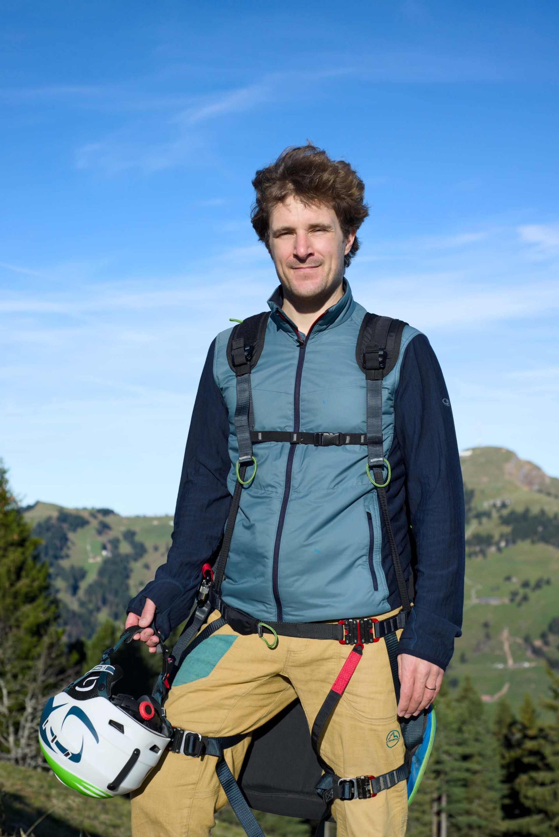 Tandem pilot Pascal Croizier stands smiling on a meadow under a blue sky with Mount Rigi in the background. He is wearing a paragliding harness and holding a white pilot helmet in his hand.