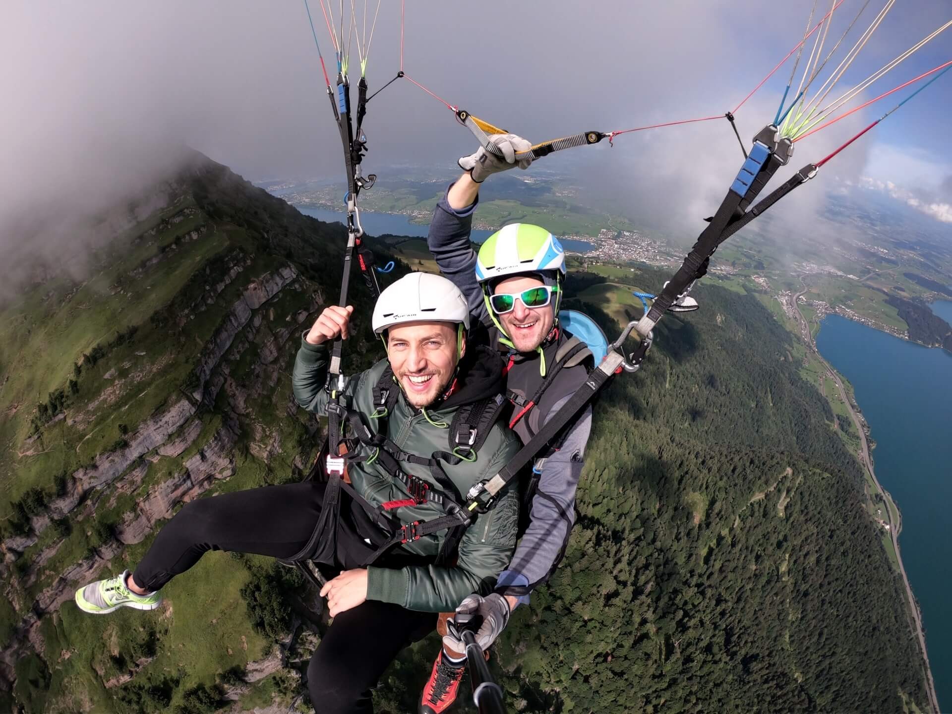 Tandem pilot Pascal flies with passenger Armand Neeskens on a tandem paraglider over the green forests of Mount Rigi. Both smile at the camera, with Lake Zug stretching out in the background.