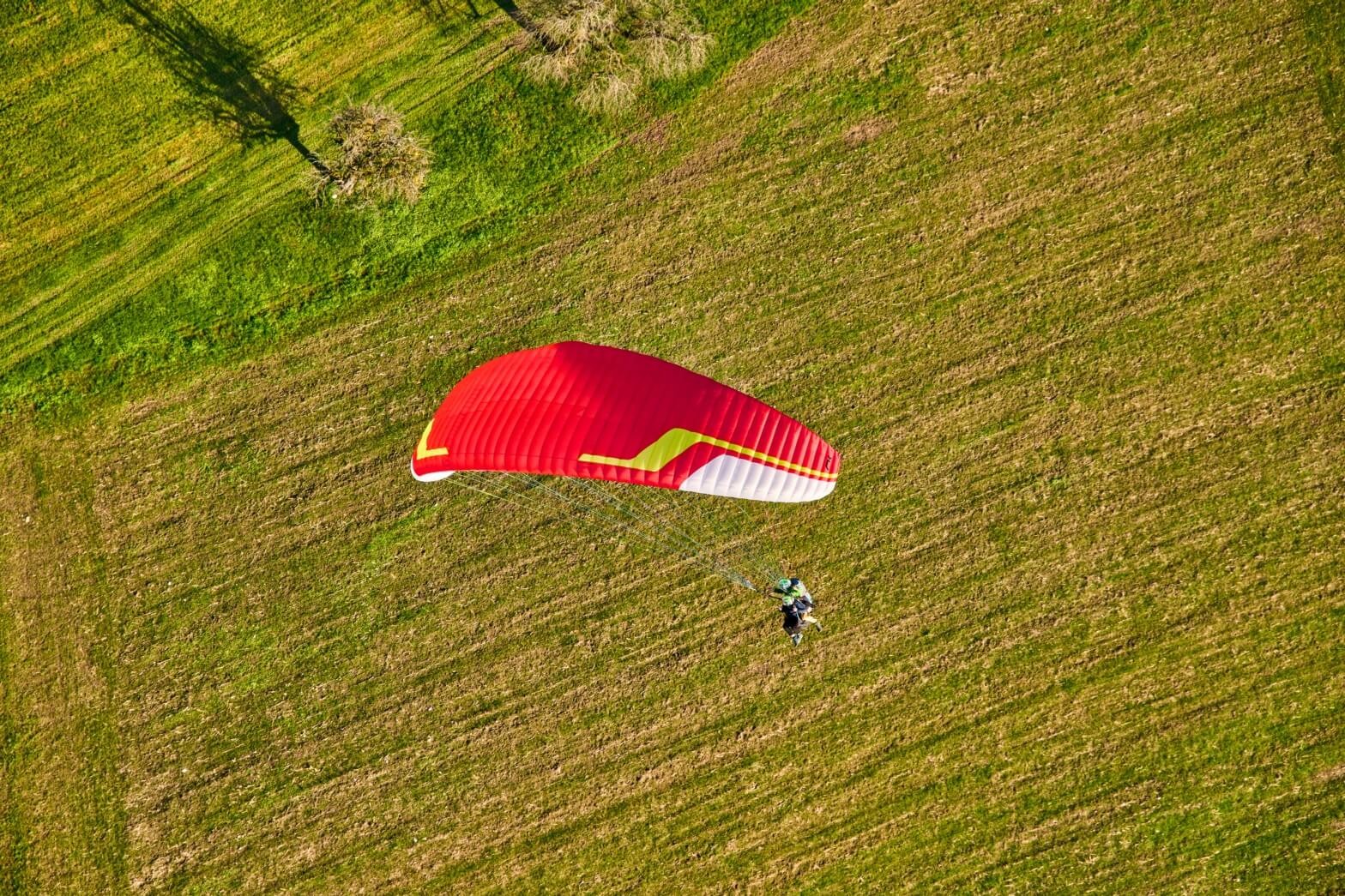 A tandem makes a turn during the landing approach over the landing site in Rickenbach near the Rotenflue. The passenger and pilot are clearly visible. A large tree provides shade on the green meadow.