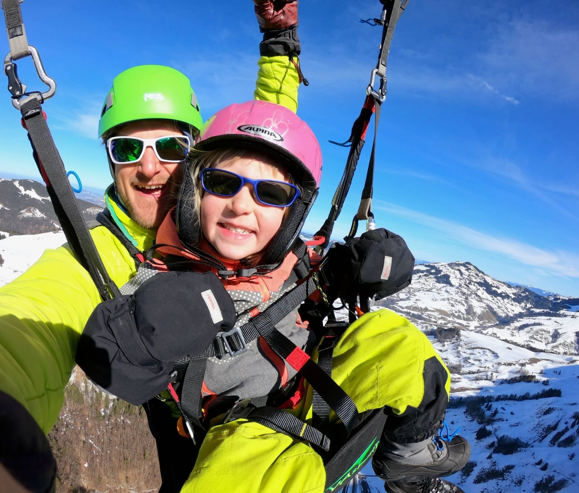 Tandem pilot Pascal Croizier flies a tandem paraglider over a snowy landscape. A small child sits comfortably in a child harness, wearing gloves and smiling cheekily at the camera.