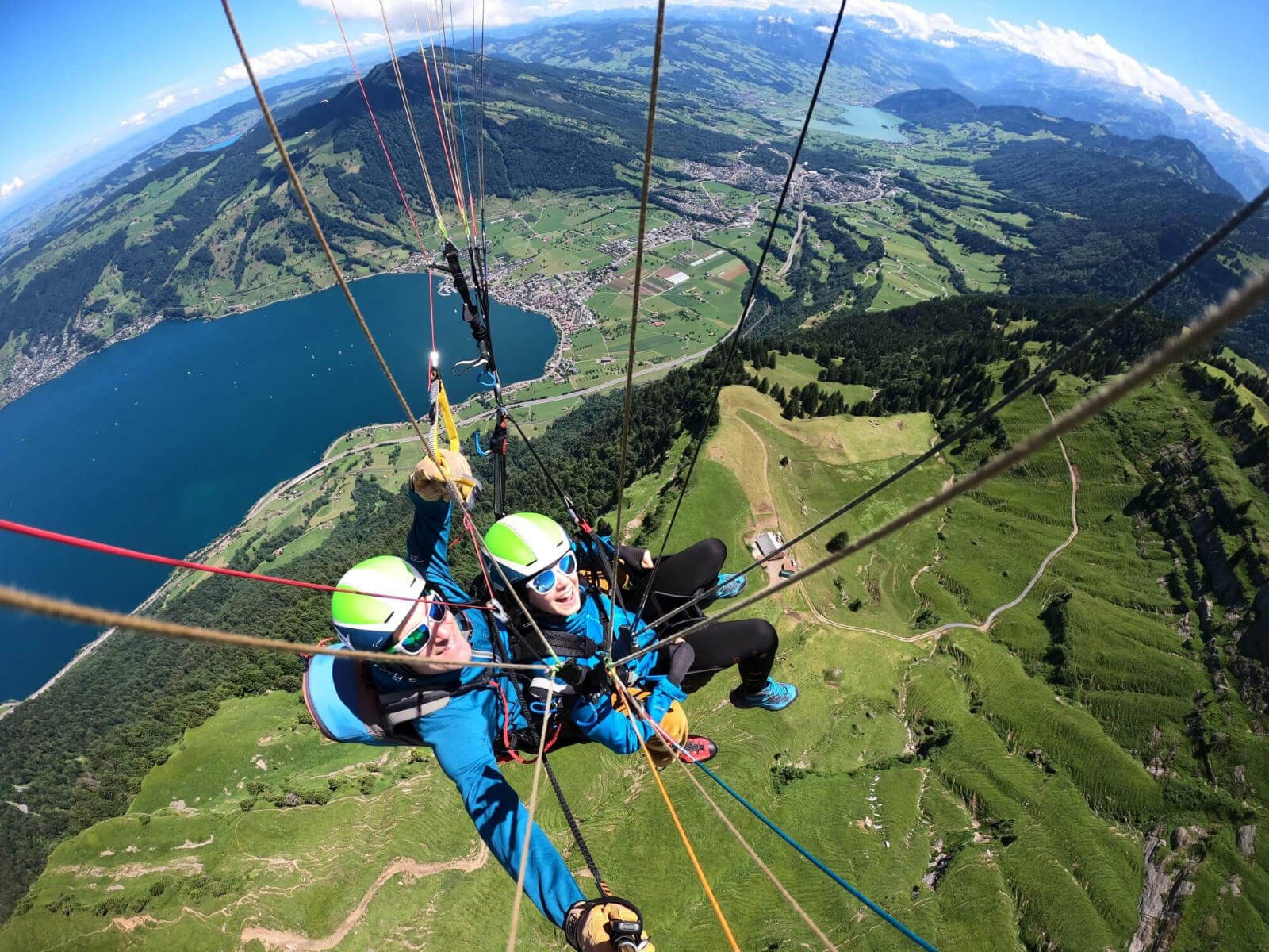 Pilot und Passagierin bei einem Tandem Gleitschirmflug über einer saftig grünen Landschaft mit Wäldern sowie der Rigi, dem Rossberg und dem Zuger- und Lauerzersee im Hintergrund. Die Passagierin lacht begeistert in die Kamera.