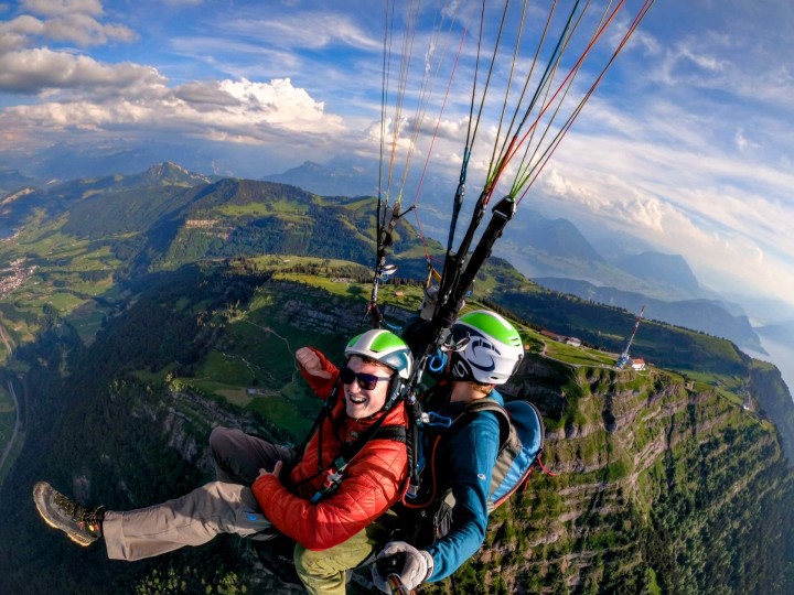 Tandemflug über der Rigi: Pilot und ein lachender Passagier genießen den Flug. Unter ihnen die steilen Felswände der Rigi, grüne Wiesen und im Hintergrund der Vierwaldstättersee mit umliegenden Bergen.