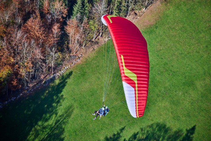 Ein Tandem macht im Landeanflug eine Kurve über den Landeplatz in Rickenbach bei der Rotenfluebahn. Passagier und Pilot sind gut zu erkennen. Ein Waldrand spendet Schatten auf der grünen Wiese.